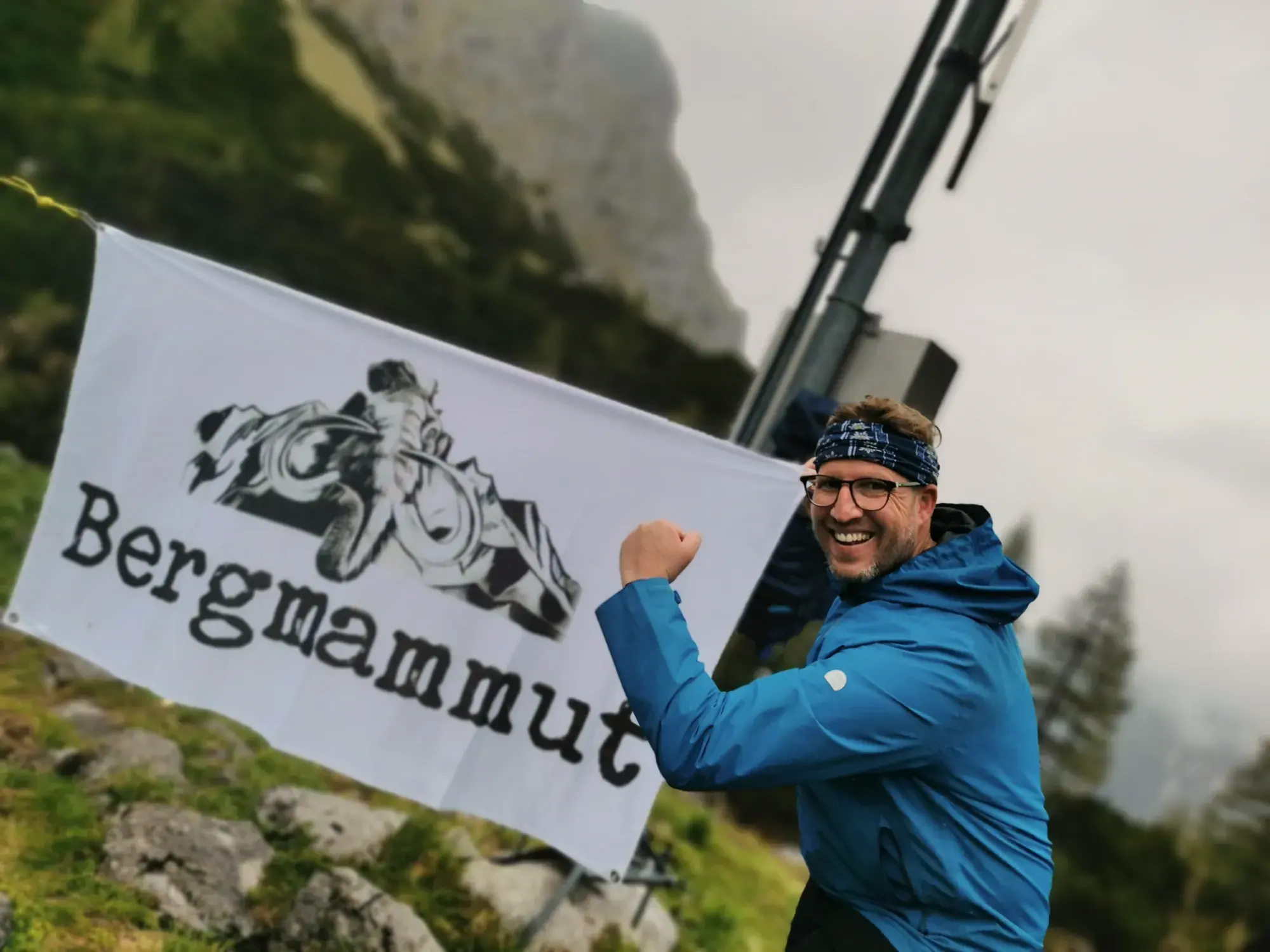 Andreas Hinz lachend neben der Bergmammut-Fahne an einem Gipfelkreuz in den Berchtesgadener Alpen.