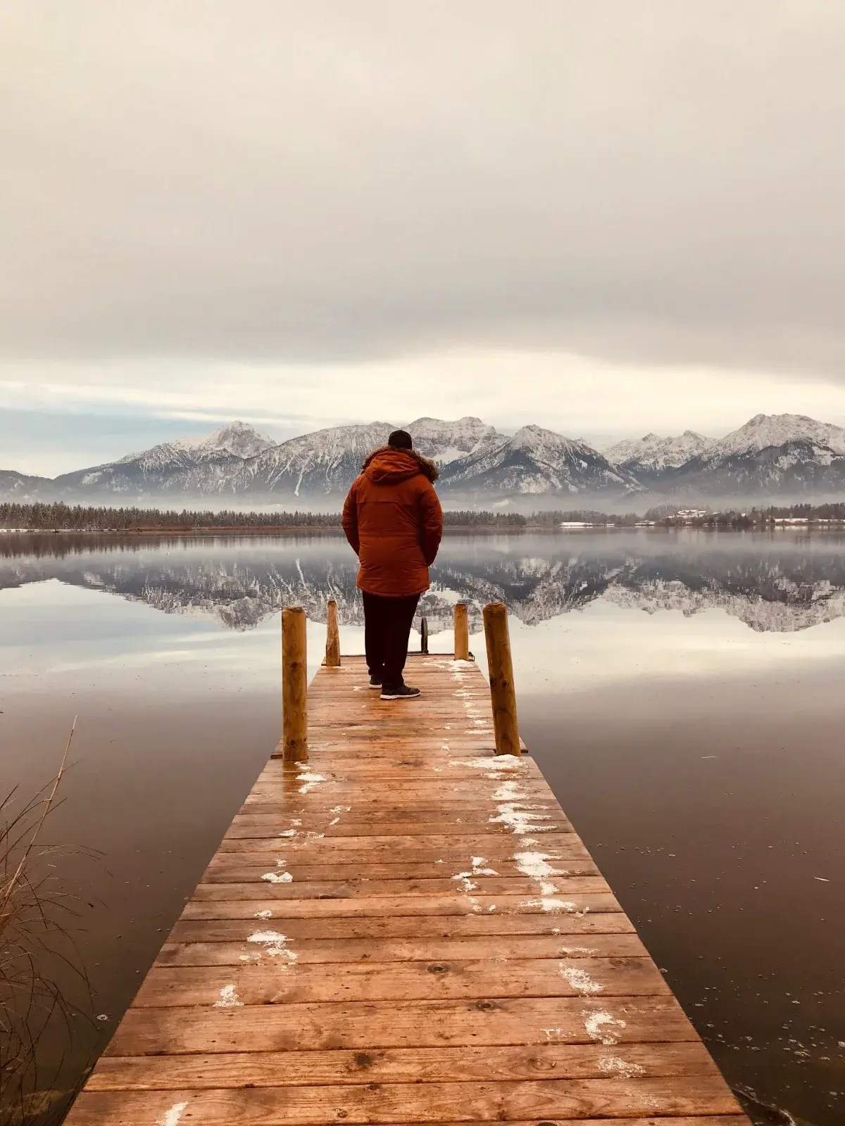 Andreas Hinz in orangefarbener Jacke am Ende eines Holzstegs am Hopfensee, im Wasser spiegeln sich verschneite Berge.